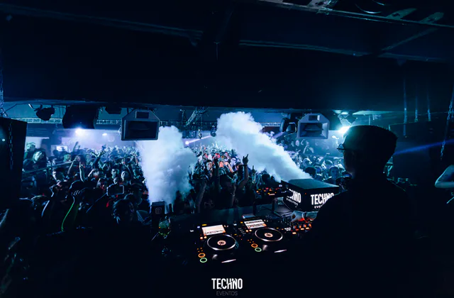 Cabina de DJ en primer plano y público reunido detrás bajo luces blancas.